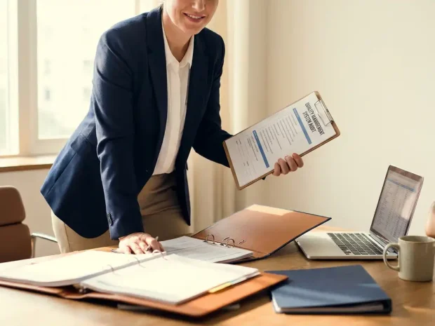 Professional in zakelijk pak bekijkt kwaliteitsmanagement checklist op klembord aan modern bureau met laptop en ordners.